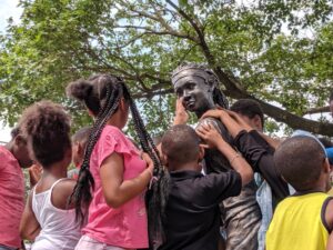 A group of Black children reach out to touch the face of a sculpture depicting a young Black female athlete.