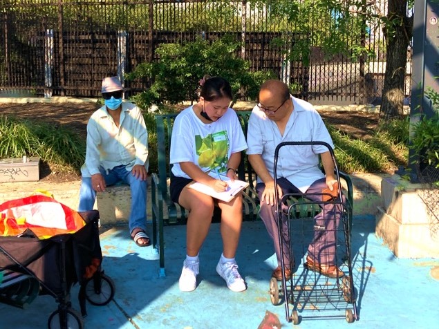 Photograph of a young woman conducting an intercept survey with a senior gentleman in Chinatown Philadelphia. They are both sitting in a planter under the trees, with a shopping cart nearby. Fotografía de una mujer joven realizando una encuesta de interceptación a un señor mayor en Chinatown Philadelphia. Ambos están sentados en una jardinera bajo los árboles, con un carrito de las compras cerca. 