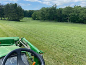 A scenic landscape perspective from the seat of a tractor over a large, grassy, fenced-in field surrounded by a forest of trees