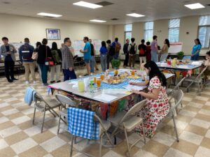 A group of people attend an Open House event about the Chinatown Stitch and a table in the center of the room has snacks like boba tea, egg tarts, and buns.