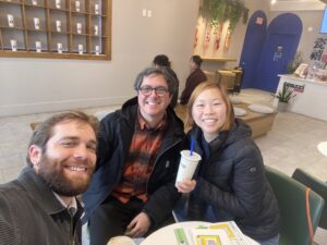 A bearded man, a man wearing glasses and an Asian woman sit together at a table and smile for a group photo