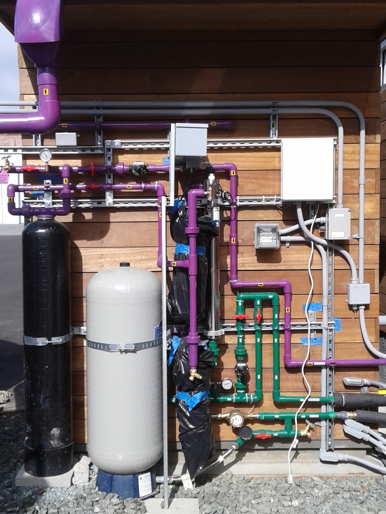 The back of a small building showing several water tanks and many pipes. 