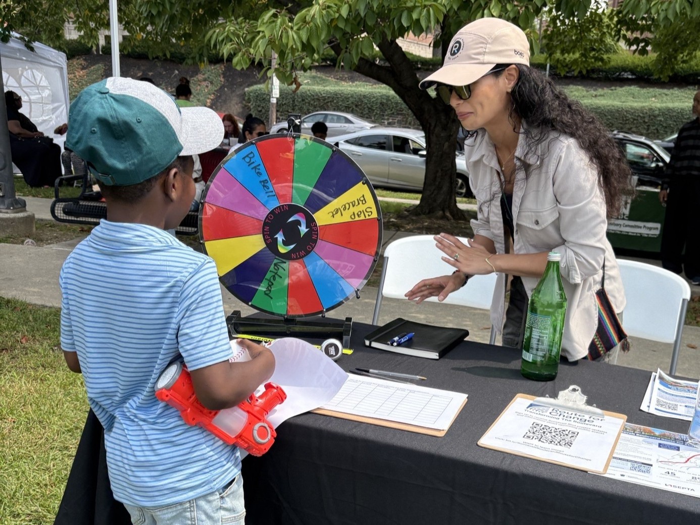 A woman wearing a tan cap leans over a table at an outdoor community event, speaking with a young boy in a green cap who is holding a red toy blaster. On the table is a colorful prize wheel, clipboards with sign-in sheets, flyers, and a green bottle. They are set up under a tent on a grassy area with trees, parked cars, and other people in the background.