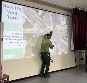 A participant stands at the front of a room, pointing to a large projected aerial map of a Philadelphia intersection during an environmental justice workshop. The slide reads, “Now It’s Your Turn: A major intersection in Philadelphia needs your help.” The participant gestures to the map while holding a paper, engaging in an interactive exercise to identify opportunities for greening and street improvements.